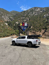 White Tacoma truck with rooftop tent (RV rental option) parked in mountains. A large, graffiti-painted structure (Pac-Man, T3K) is visible.