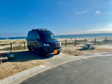 Coastal camping with an RV rental: A dark camper van parked on a concrete pad with beach access, ocean views, and a picnic table.