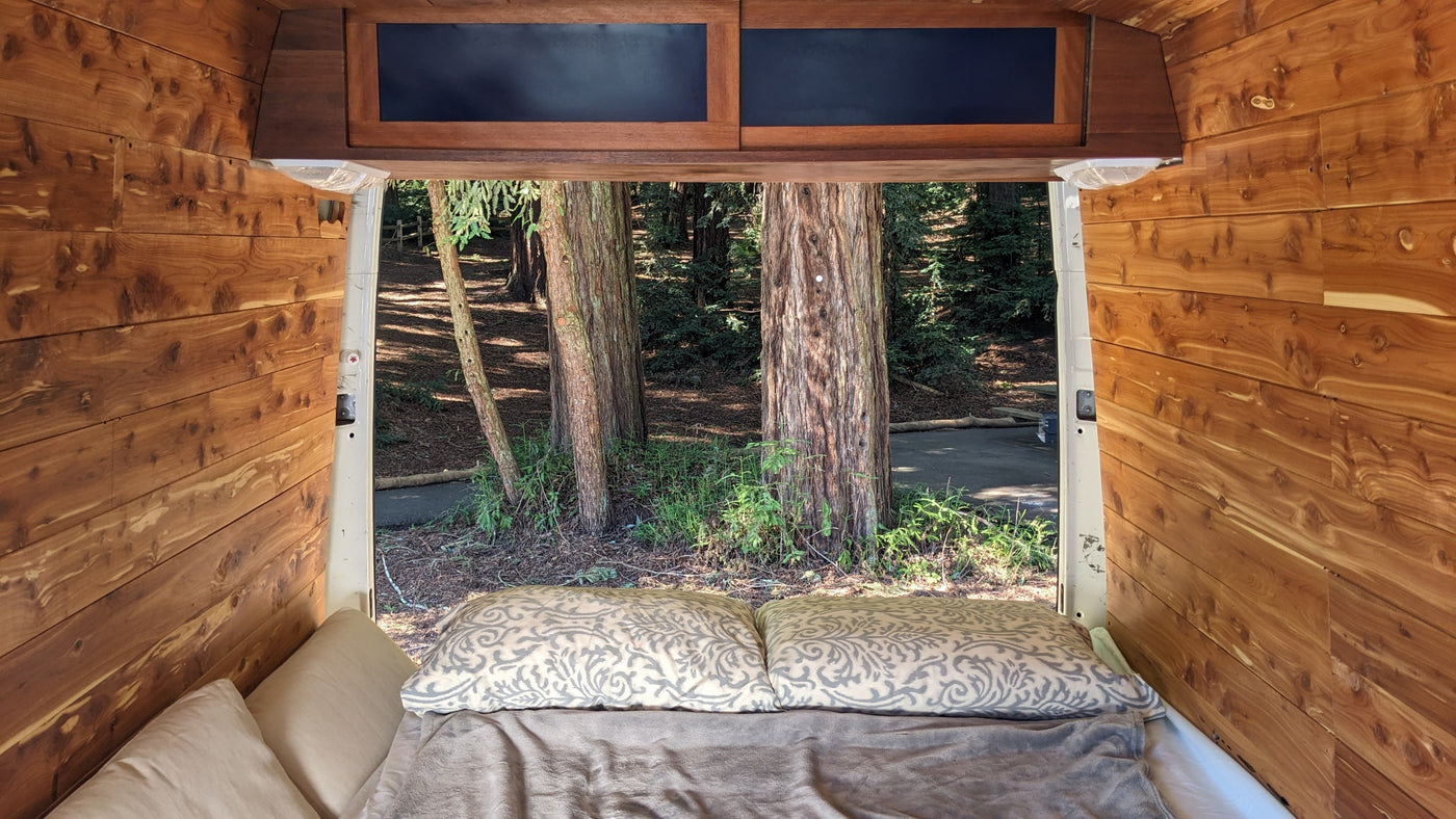Cozy rv rental camper interior, looking out the open back to a serene forest with large trees, wood-paneled walls, and a comfy bed.