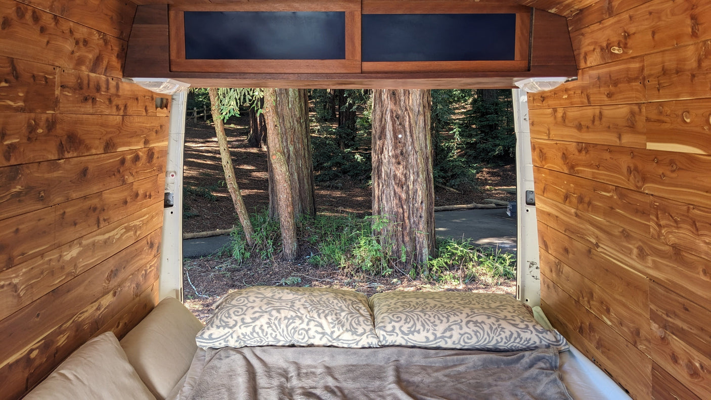 Cozy rv rental camper interior, looking out the open back to a serene forest with large trees, wood-paneled walls, and a comfy bed.