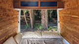 Cozy rv rental camper interior, looking out the open back to a serene forest with large trees, wood-paneled walls, and a comfy bed.