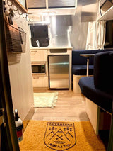 Cozy Airstream RV rental interior with compact kitchen, dining, and "Adventure Awaits" mat on the floor.