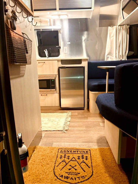 Cozy Airstream RV rental interior with compact kitchen, dining, and "Adventure Awaits" mat on the floor.