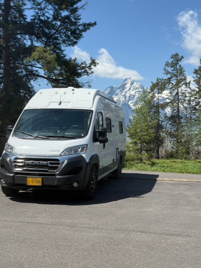 White Winnebago camper van rv rental parked against a backdrop of majestic, snow-capped mountains & pine trees.