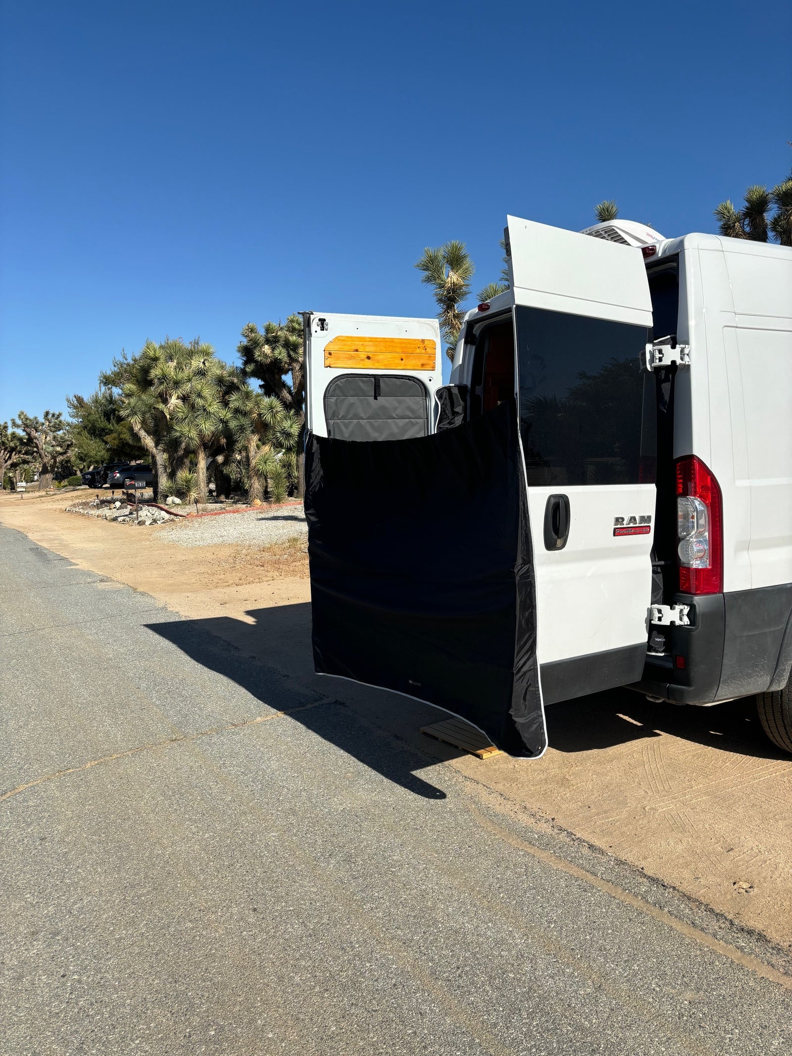 An RV rental van's open rear doors with a privacy screen set up, amidst Joshua trees on a sunny day.