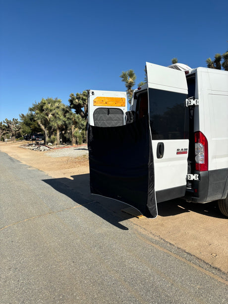 An RV rental van's open rear doors with a privacy screen set up, amidst Joshua trees on a sunny day.