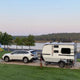 Explore the outdoors! This small travel trailer, possibly an RV rental, is perfectly towed by a white car at a lakeside campsite.