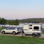 Explore the outdoors! This small travel trailer, possibly an RV rental, is perfectly towed by a white car at a lakeside campsite. — photo 2