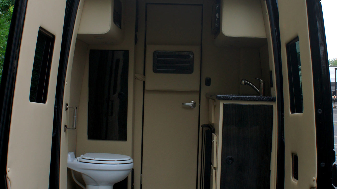 Inside an RV rental, a compact interior features a white toilet on the left and a kitchenette with a sink on the right.