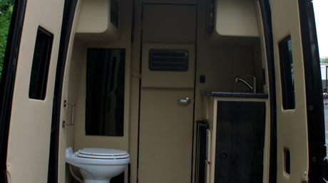 Inside an RV rental, a compact interior features a white toilet on the left and a kitchenette with a sink on the right.
