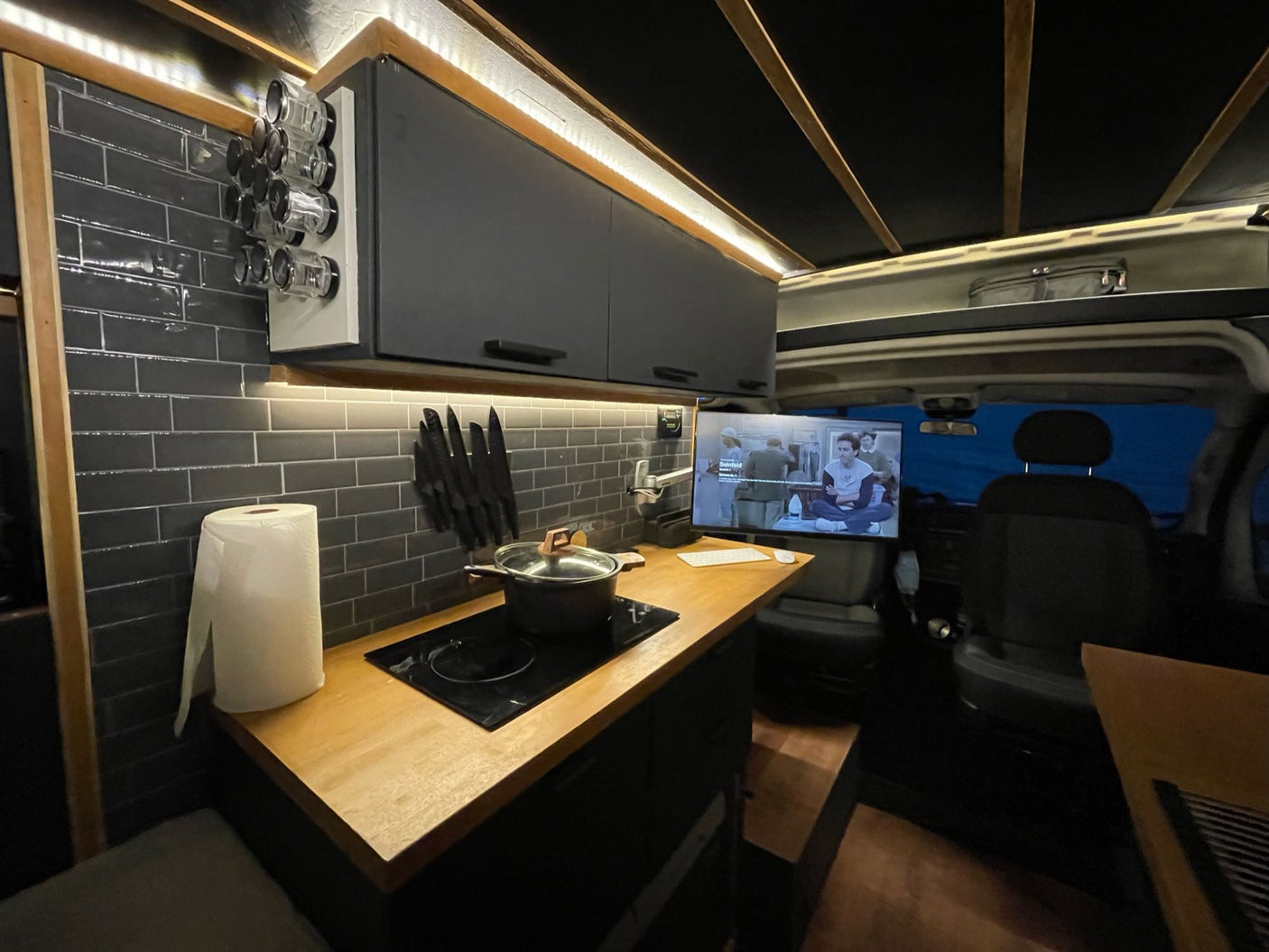 This modern RV rental kitchen features dark tiled walls, wood counters, an induction cooktop, spice rack, and TV.