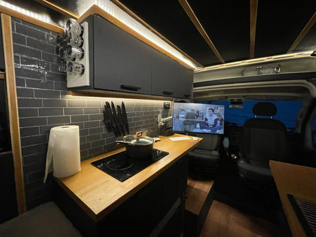 This modern RV rental kitchen features dark tiled walls, wood counters, an induction cooktop, spice rack, and TV.