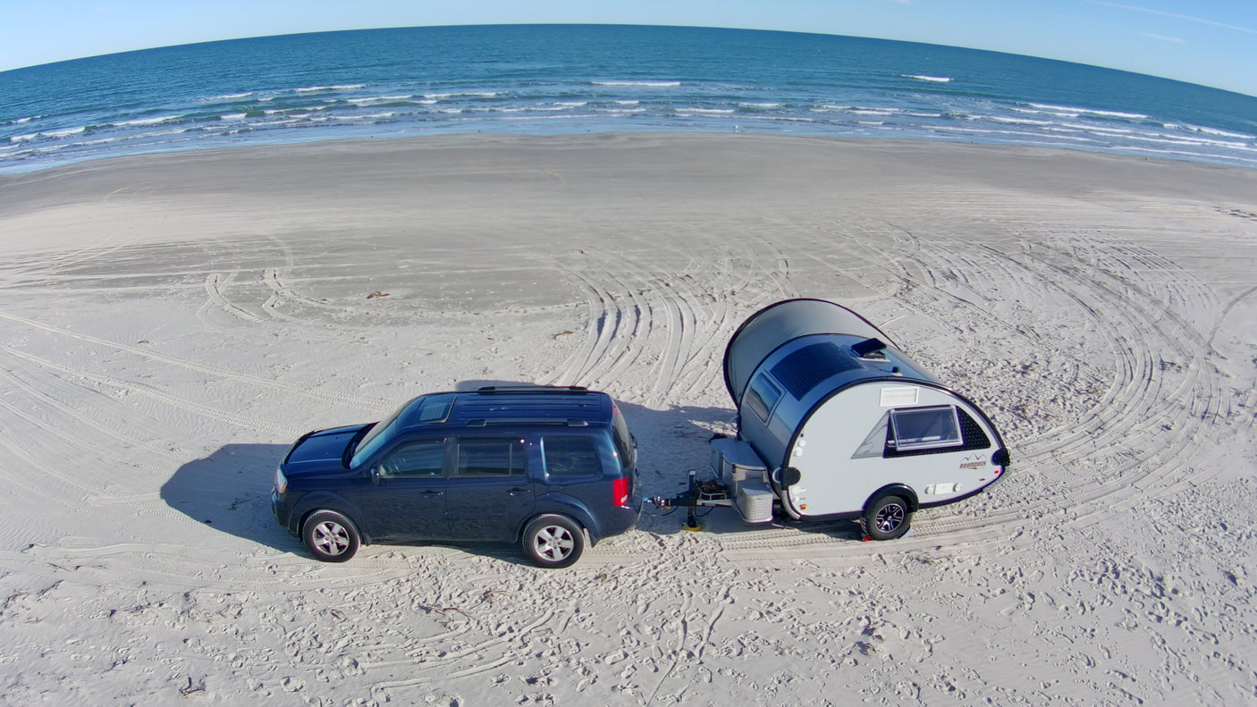 A dark blue SUV tows a gray teardrop RV rental camper on a wide sandy beach, with ocean waves visible in the background.