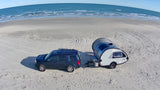 A dark blue SUV tows a gray teardrop RV rental camper on a wide sandy beach, with ocean waves visible in the background.