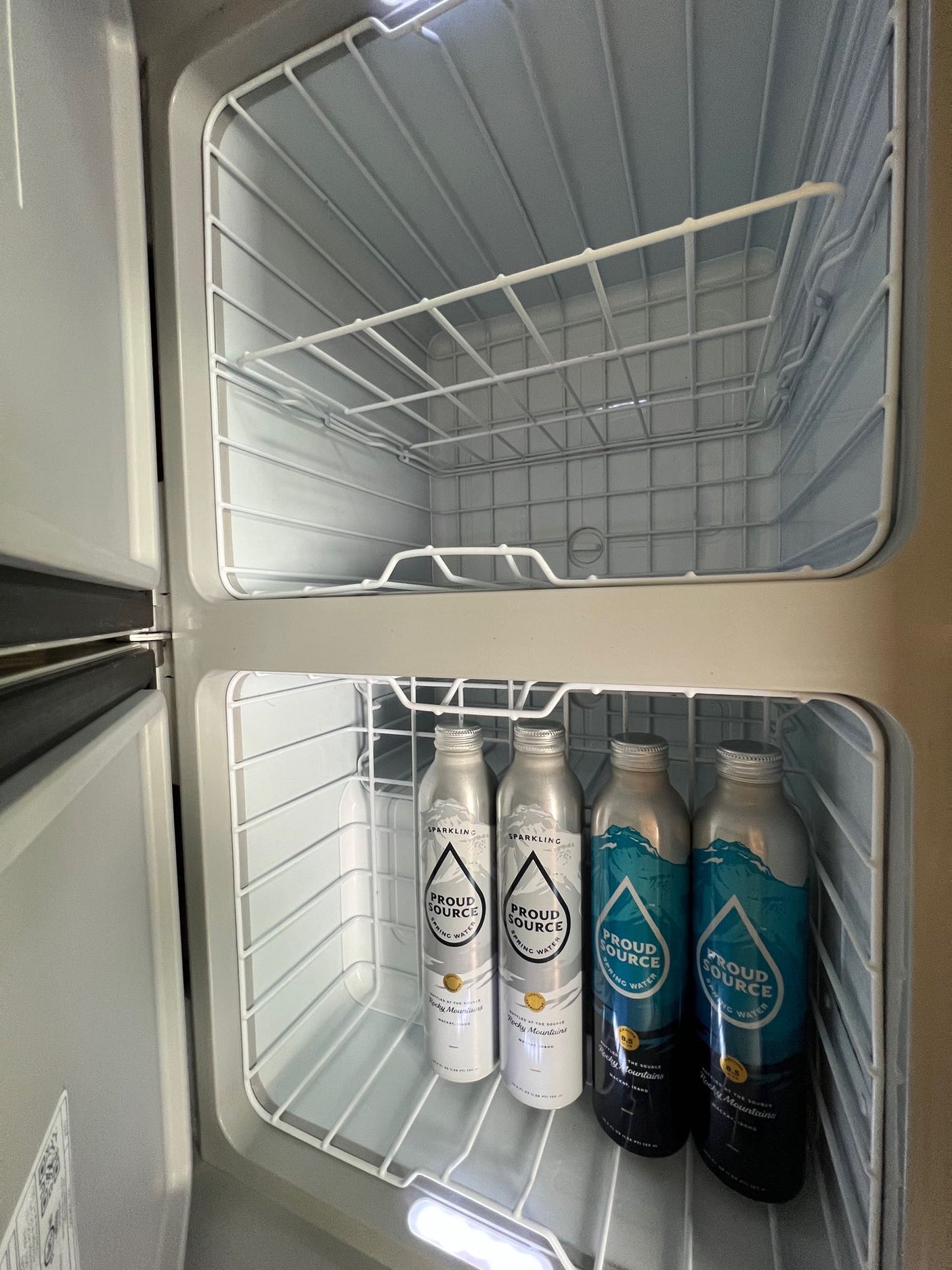 Inside an RV rental compact fridge: empty top compartment and Proud Source sparkling/spring water bottles on the lower shelf.