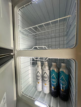 Inside an RV rental compact fridge: empty top compartment and Proud Source sparkling/spring water bottles on the lower shelf.