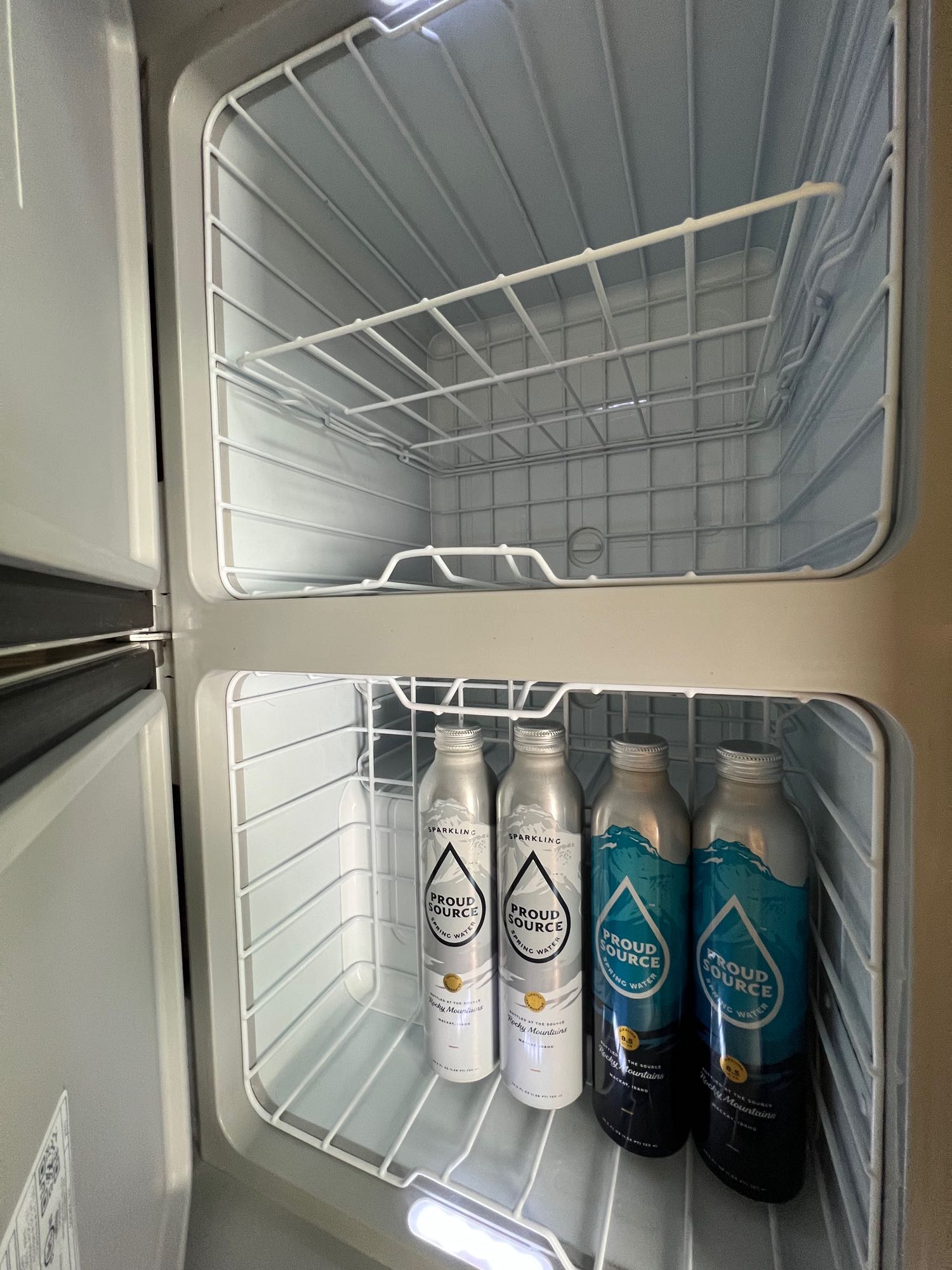 Inside an RV rental compact fridge: empty top compartment and Proud Source sparkling/spring water bottles on the lower shelf.