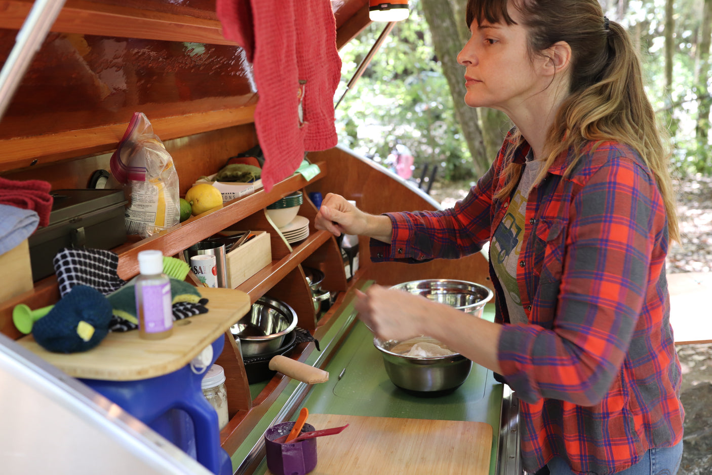 A woman in a plaid shirt mixes food in her RV rental's outdoor kitchen, enjoying campsite cooking amidst nature.