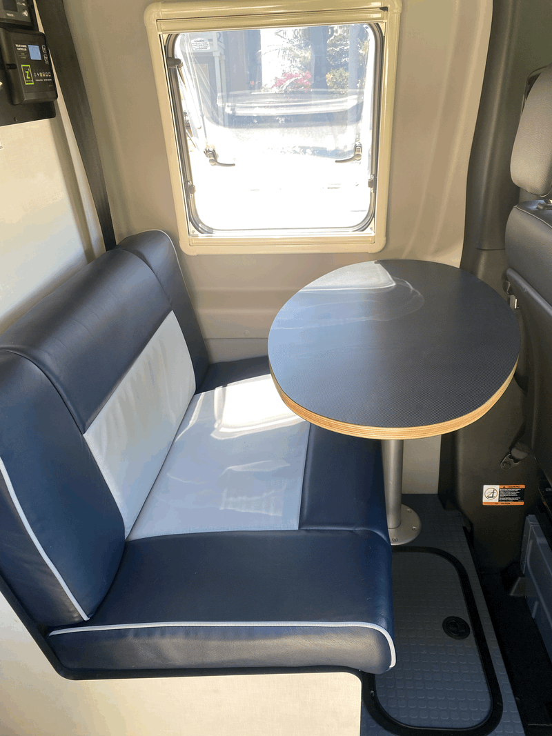Interior view of an RV rental dinette area with blue and white bench seating, a round table, and a window. Features a solar charge controller. — photo 11
