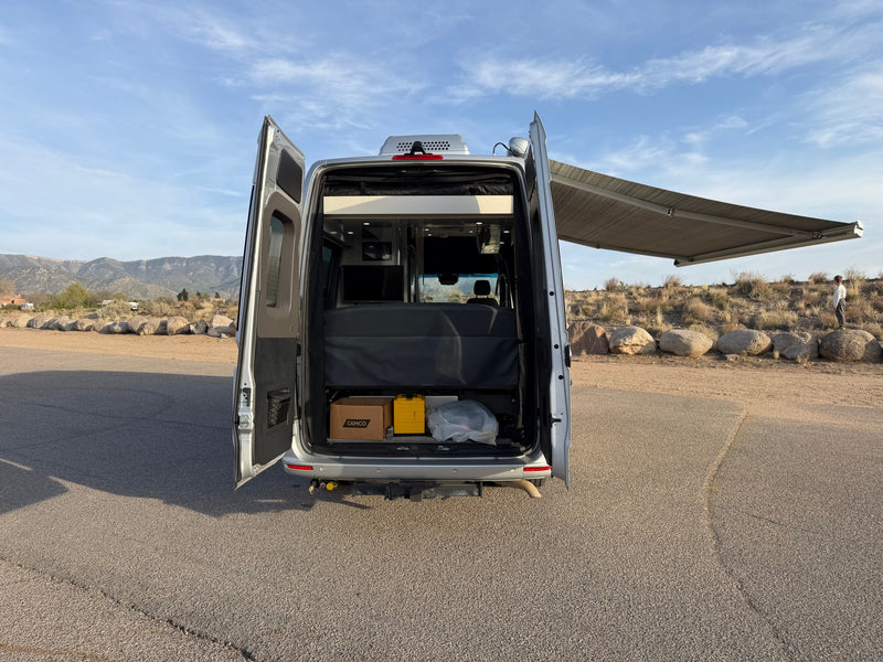 Open back of a silver camper van RV rental, showing interior storage, bed, extended awning, and scenic views. — photo 19