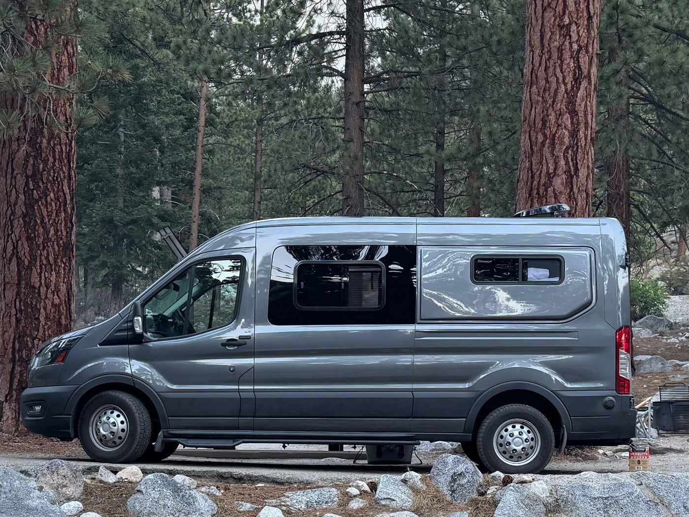 Grey rv rental campervan parked sideways amidst tall pine trees at a rocky forest campsite.|An RV rental camper van parked amidst desert brush with mountains glowing orange at golden hour. Scenic view.|An RV rental camper van parked at a serene forest campsite. Picnic table — photo 1