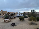 An RV rental van at a rugged desert campsite with a picnic table, fire ring, rocks, and Joshua trees under a cloudy sky.