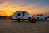 This RV rental: a white teardrop camper and silver truck parked on a sandy beach at sunset, ideal for camping.
