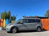 A gray RV rental van with a surfboard on its roof, parked outdoors with colorful surfboards and bamboo in the background.