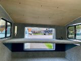 Inside an RV rental: a cozy space with carpeted walls, wooden ceiling, power strip, and scenic views from multiple windows.