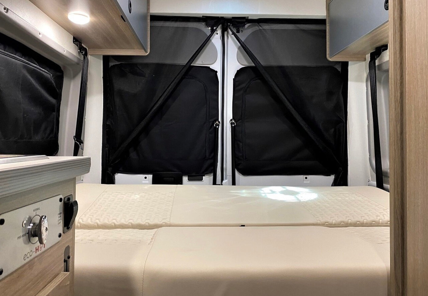 RV rental interior: A cozy bed faces rear doors with black window covers. A control panel, "eco-Hot," is on the left.
