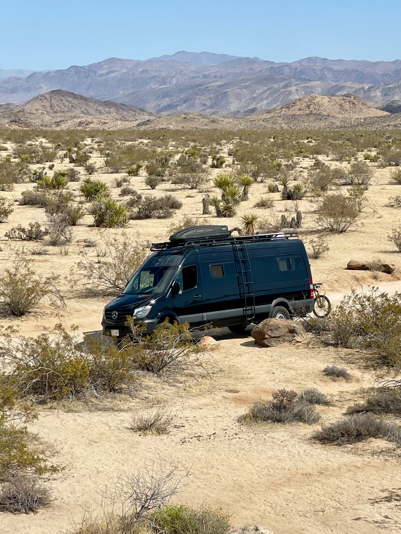 Adventure awaits! Dark RV rental van with bike in a rugged desert landscape with mountains.