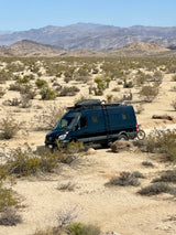 Adventure awaits! Dark RV rental van with bike in a rugged desert landscape with mountains.