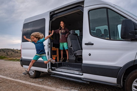 Two excited children leap from a white RV rental van, embracing outdoor adventure.