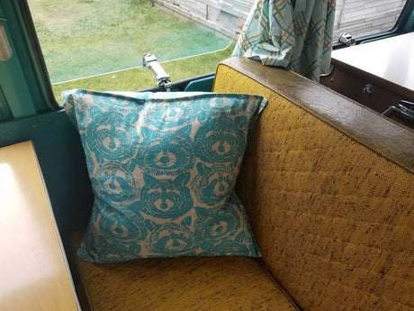 Inside an RV rental, a cute teal raccoon pillow rests on a mustard yellow patterned bench seat, with a view outside.
