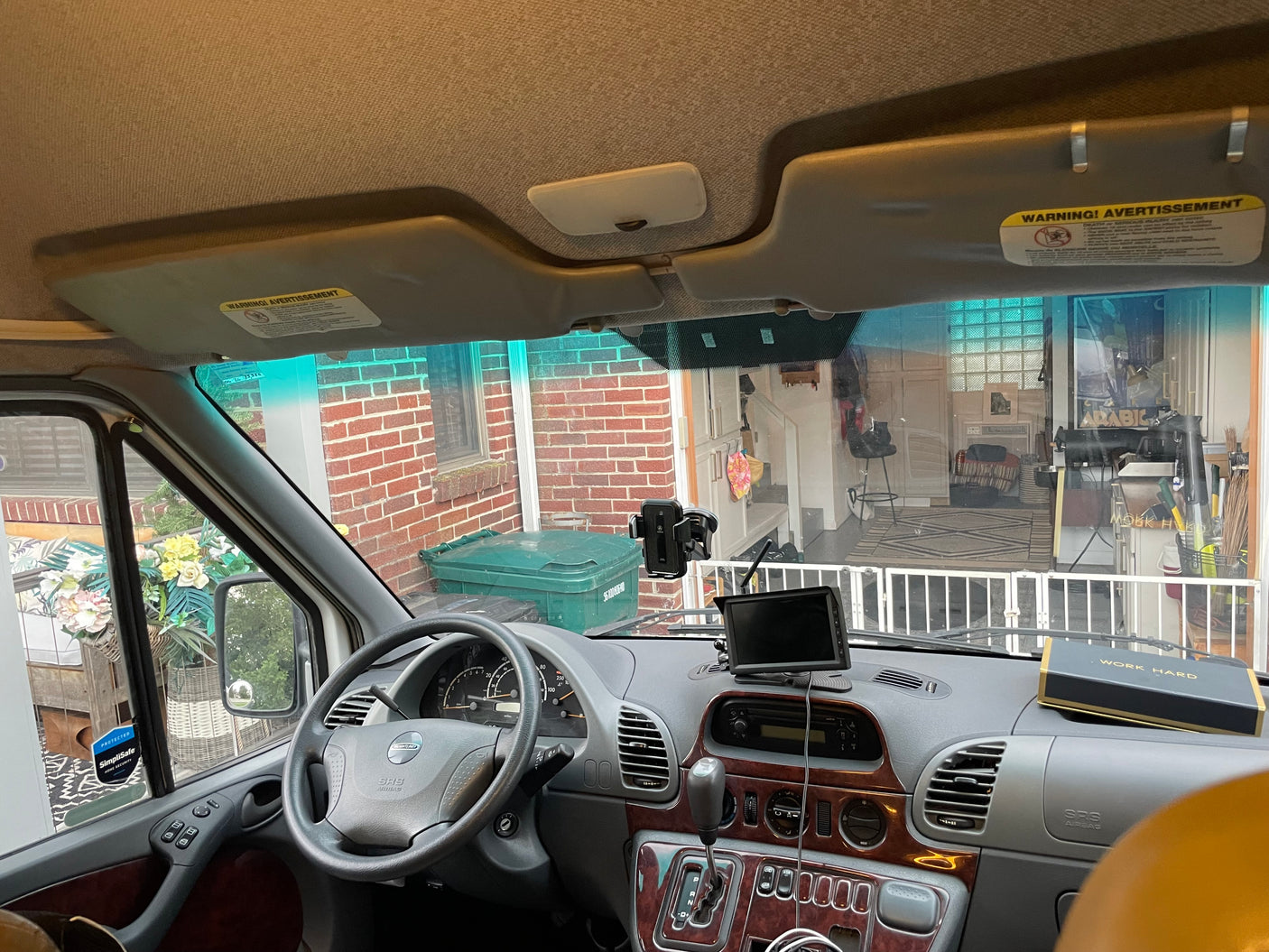RV rental interior: Driver's view of the dashboard, steering wheel, and the exterior through the windshield towards a brick house.