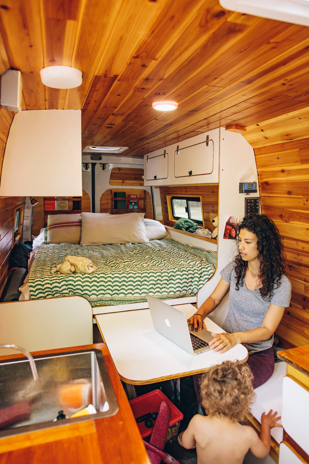 Van life in an RV rental: Woman on laptop, child playing, inside a compact camper van with a bed and kitchen.