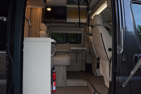 Interior of an rv rental from the open side door: shows a kitchenette, seating area with table, TV, and front driver seats.