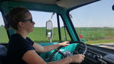 Inside a vintage RV rental: a woman drives the teal vehicle past green fields, wind turbines visible.