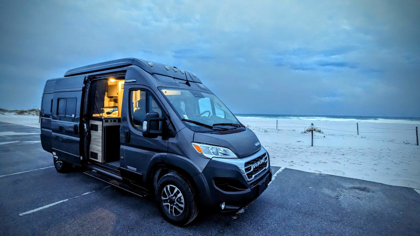 Dark grey RV rental parked by a sandy beach at dusk, interior lit. Ideal for beach travel and adventure.