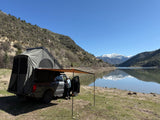 A truck with rooftop tent and awning by a scenic lake, with snow-capped mountains reflected. A perfect outdoor getaway, like an RV rental.