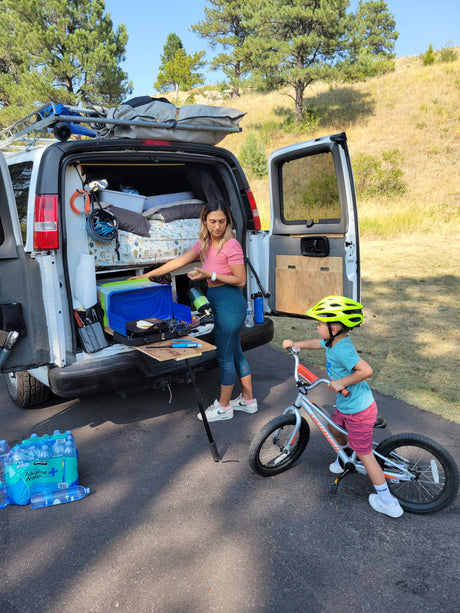 RV rental camp setup: a woman cooks from the back of a van, a child cycles nearby. Features a portable kitchen & bed.