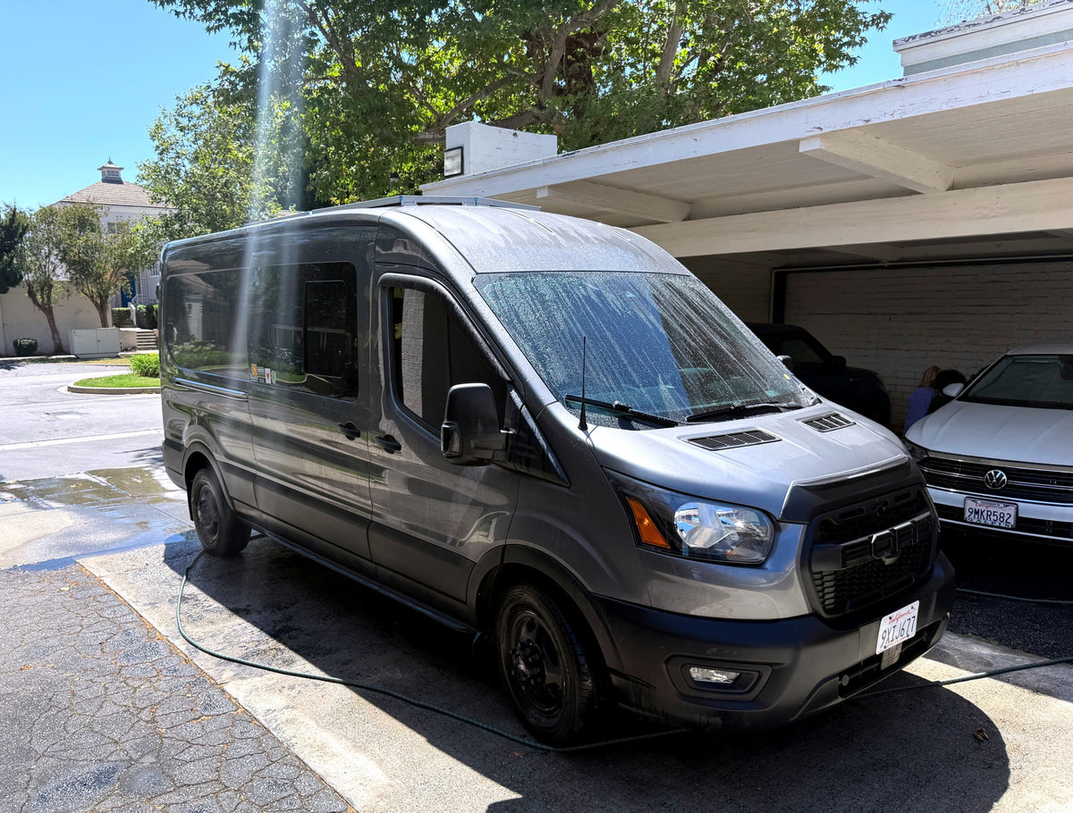 Gray Ford Transit van, likely an RV rental, being washed with a hose in a sunny driveway partly under a carport.