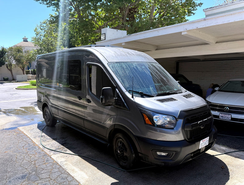 Gray Ford Transit van, likely an RV rental, being washed with a hose in a sunny driveway partly under a carport. — photo 1