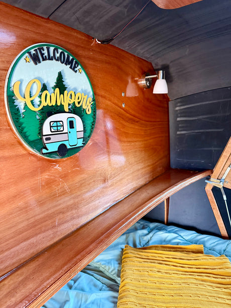 Cozy rv rental interior featuring a "WELCOME Campers" sign on wood paneling, a small light, and a comfy bed.