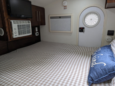 Cozy RV rental interior with a bed, checkered blanket, TV, AC unit, storage, window, and a unique round door.