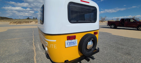 Rear view of a yellow & white P.O.P. Lite rv rental with a Michigan plate, parked by sand dunes.