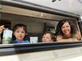 Smiling family in their RV rental window. Two boys and a woman enjoy their camping trip, one holding a lantern.