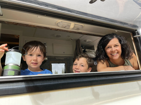 Smiling family in their RV rental window. Two boys and a woman enjoy their camping trip, one holding a lantern.
