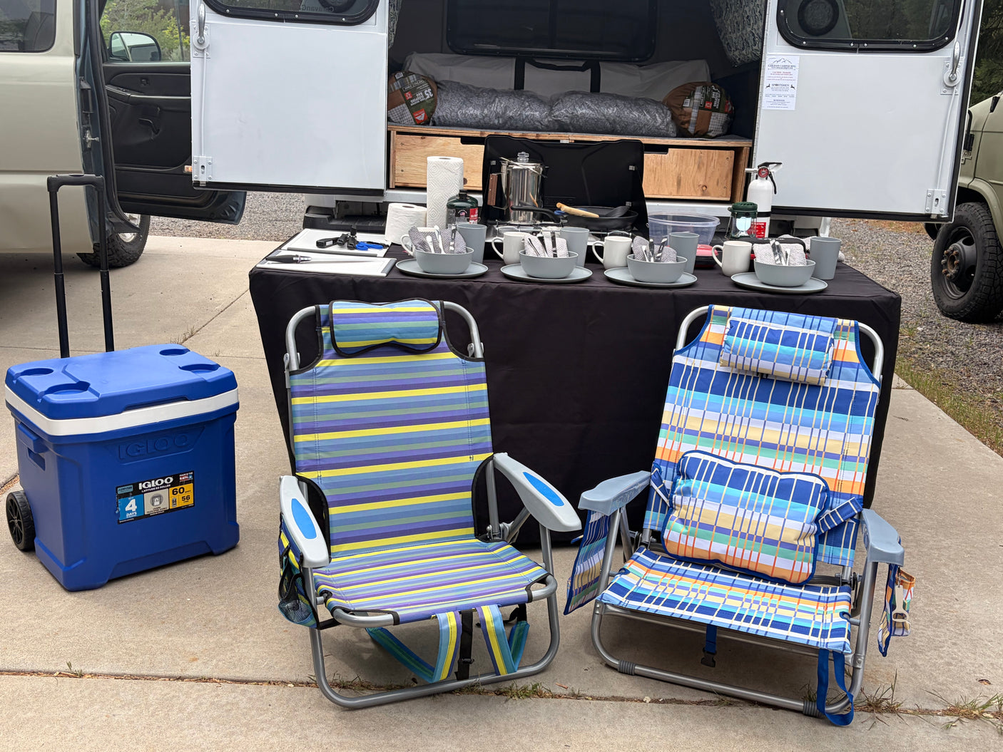 An RV rental camper, doors open, alongside a full outdoor kitchen setup, cooler, and two striped camping chairs.
