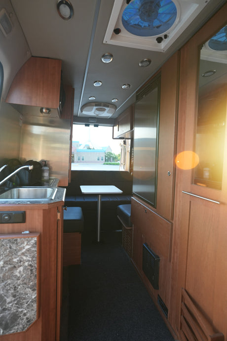 RV rental interior: modern kitchen, dining area with black benches, wood cabinetry, and ceiling fan, viewed from front.